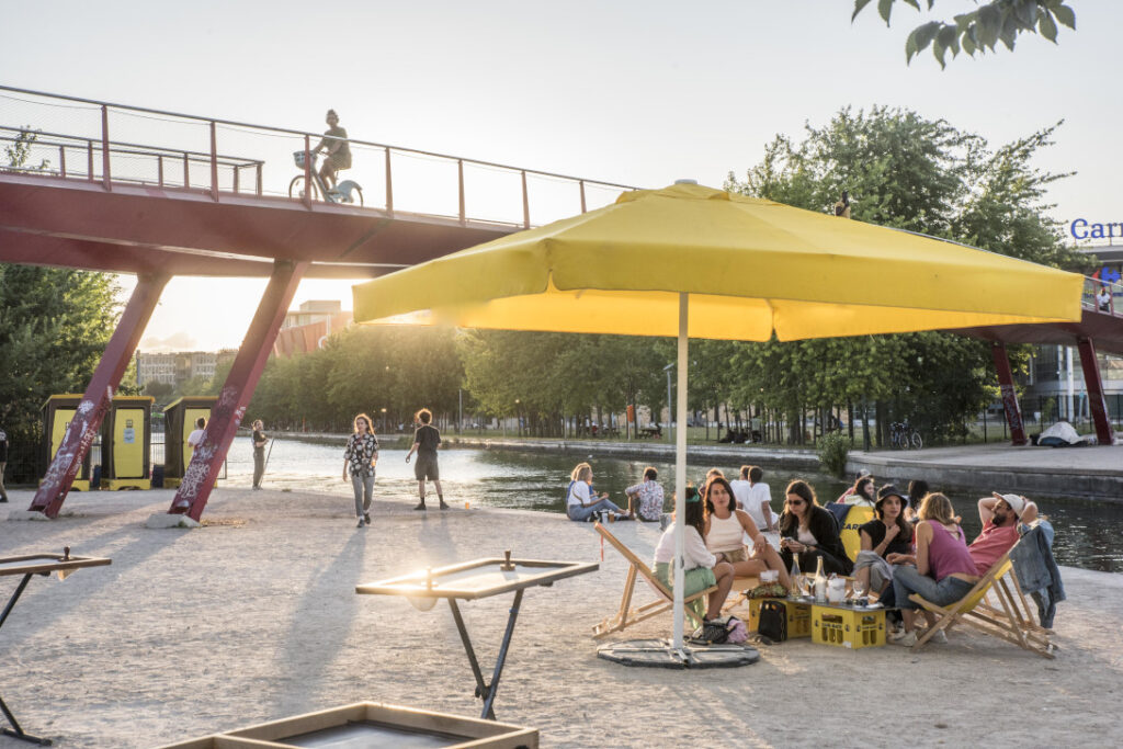 Photo de la terrasse de la péniche le Barboteur sur la place du Millenaire, montrant des personnes prenant un verre sous un parasol au bord du canal Crédit : JM Sicot