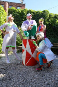 Quatre artistes en costumes colorés posent autour d’un tonneau rouge dans une cour végétalisée.